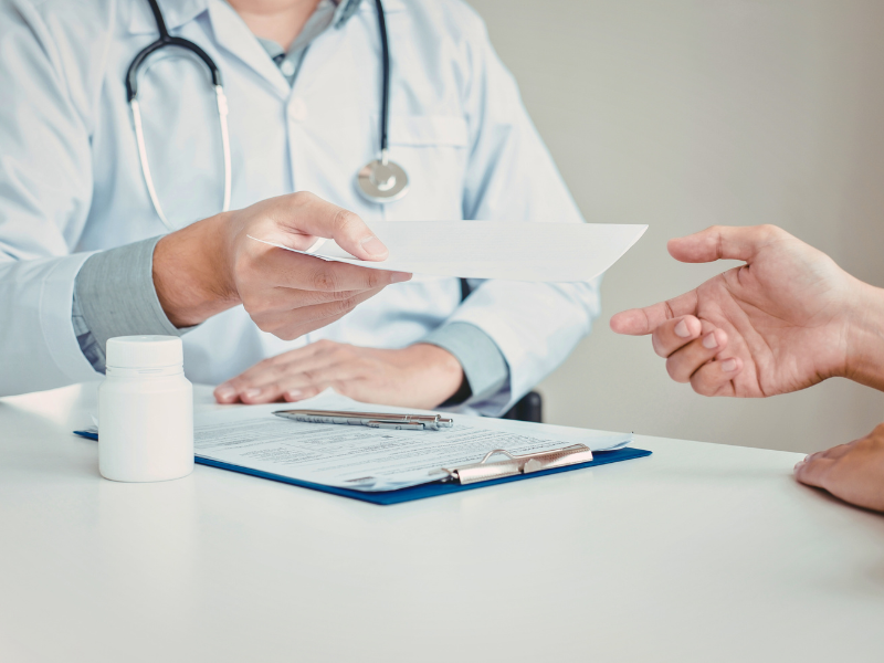 Doctor in a white coat handing a paper to a patient across a desk with a pill bottle 