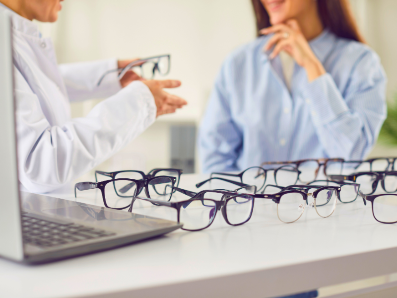 Optician presenting eyeglasses to a customer, with multiple eyeglass frames arranged on a white table 